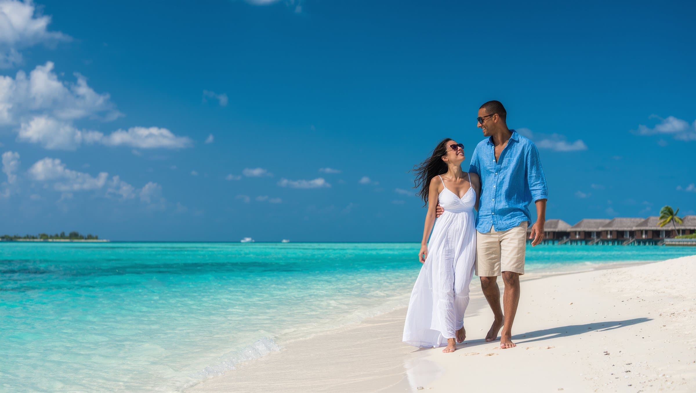 A couple walking along a white beach beside a turquoise lagoon, with over-water villas behind them